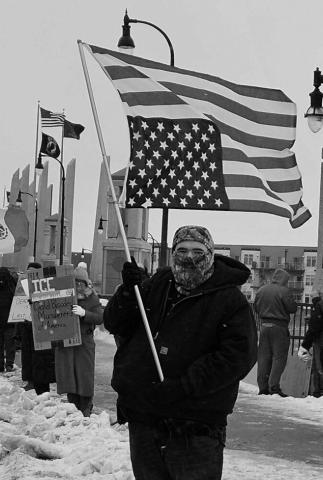 Jerrod Baetsch of West Fargo, North Dakota, stands with an American flag upside down, a sign of distress, on the Veterans Memorial Bridge over the Red River in Fargo on Jan. 10, 2026. Photo by Jeff Beach/North Dakota Monitor